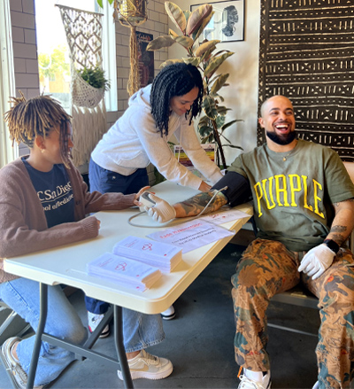 two female students conducting a blood pressure check on a male seated at a table