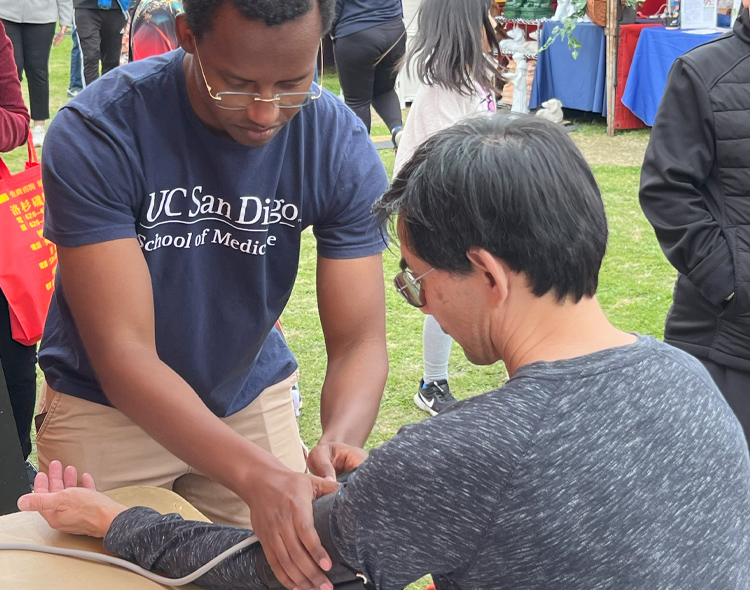 Male medical student putting a blood pressure cuff on the arm of a man sitting at a table.