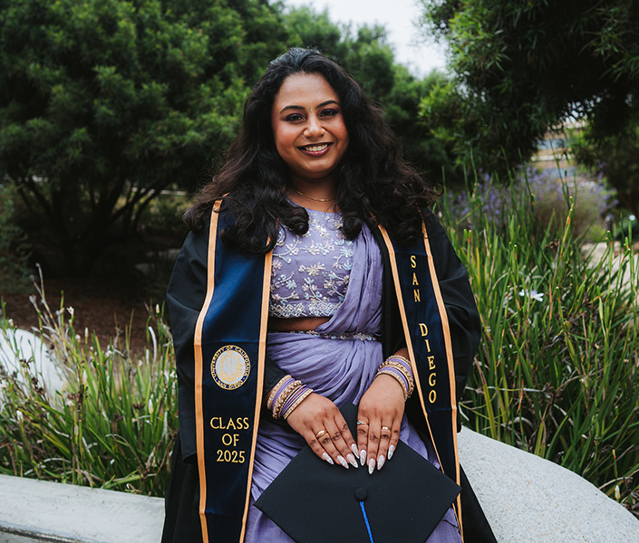 A student in commencement regalia with traditional Bangladeshi garments worn underneath. She is sitting on a rock in front of a pond.