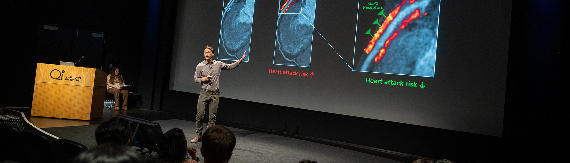 Person on stage points to slide on screen while speaking