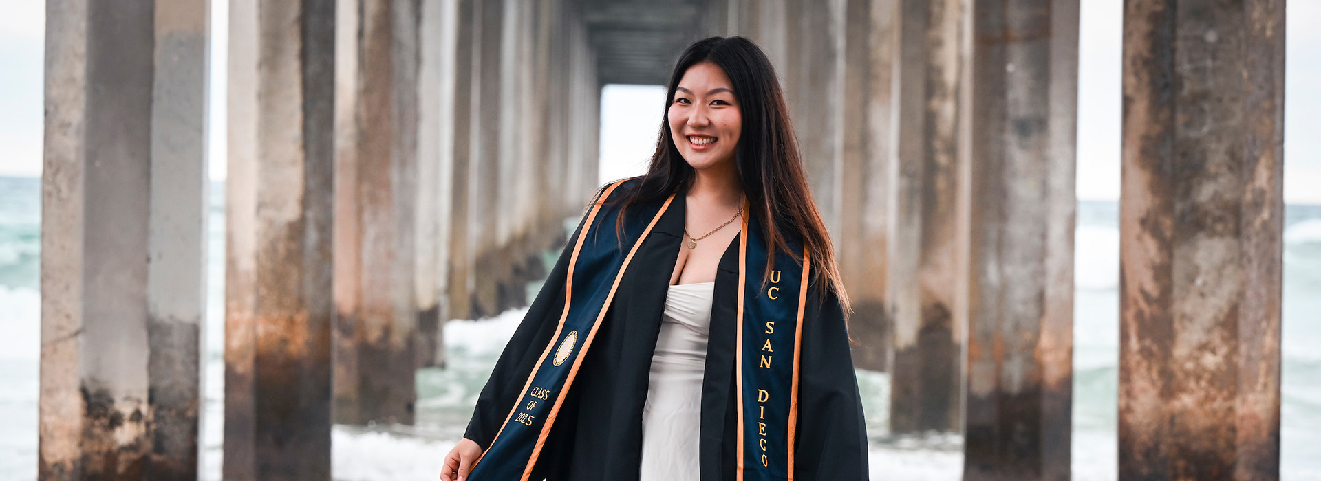 A student in commencement regalia smiling in front of a pier and the ocean.