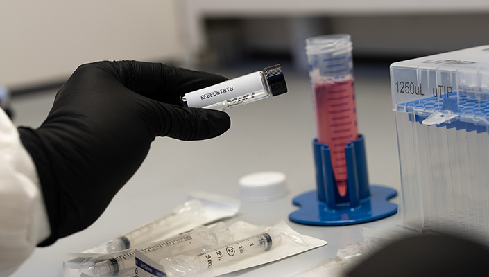 A hand in a black lab glove holds a vial of an investigational new drug.