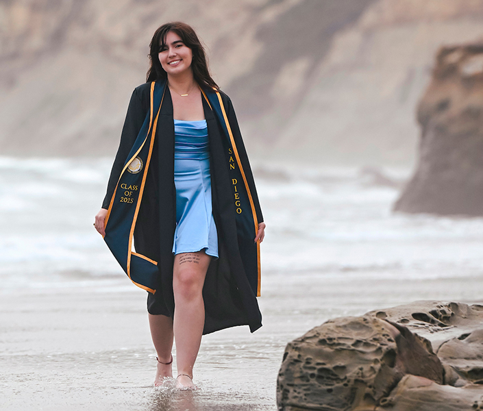 A student in commencement regalia walking on the beach shore.