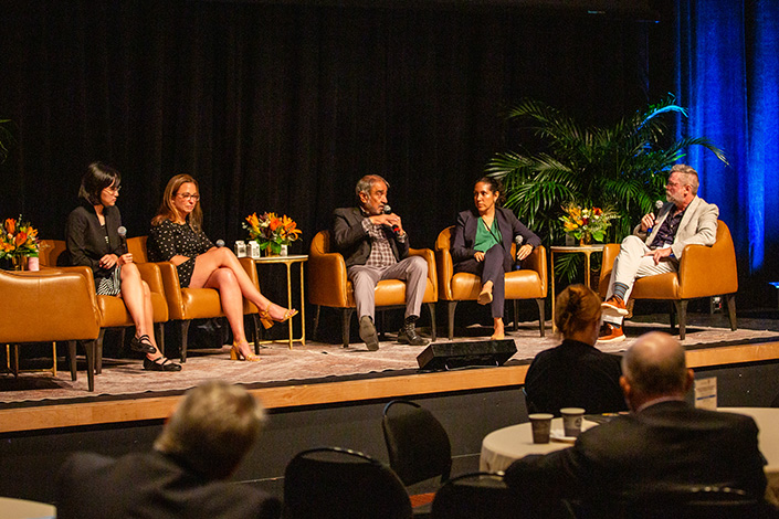 Five speakers sit in chairs on stage as audience looks on.