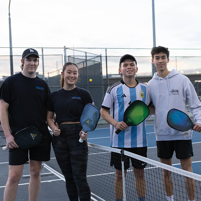 Four students hold pickleball paddles on either side of net