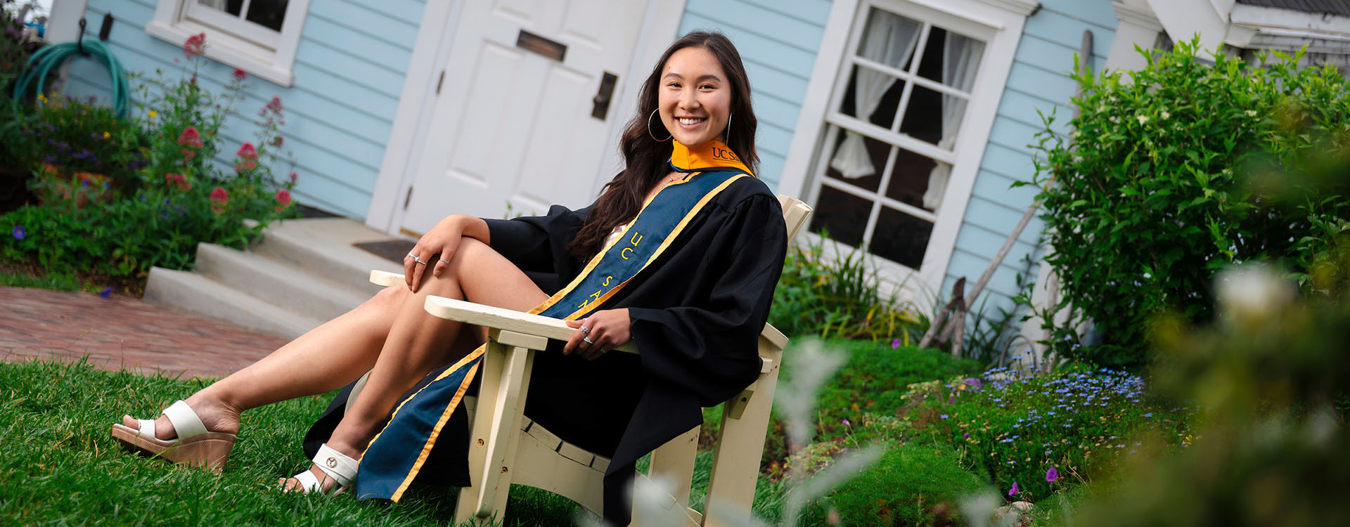 A student in commencement regalia sits in a chair on a lawn in front of a public artwork entitled Falling Star.