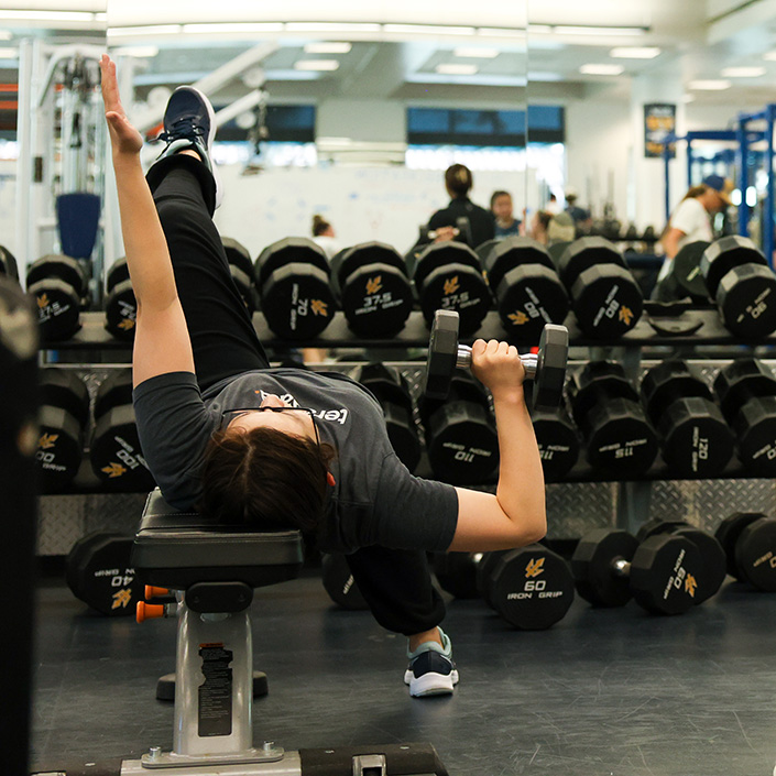 Student on weight bench holds a dumbbell in right hand and holds left hand and leg in the air