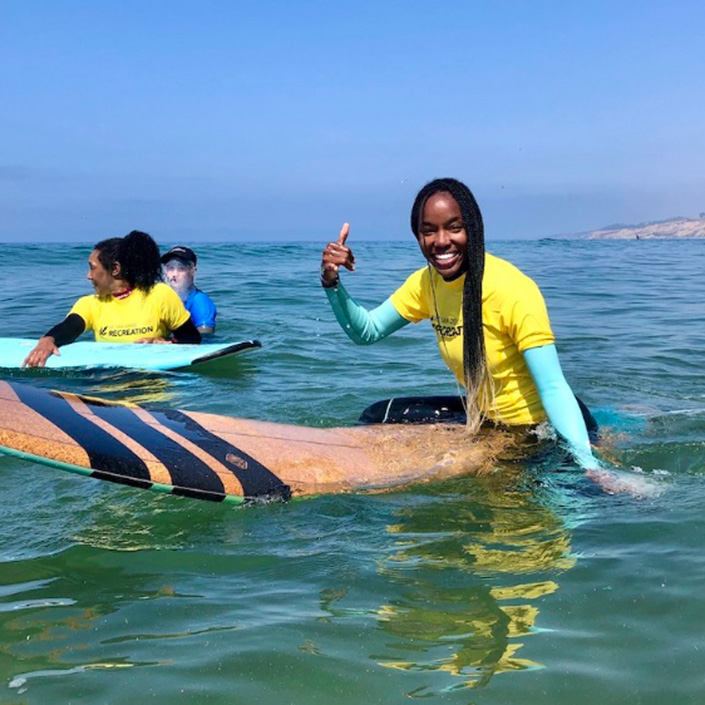 Student does shaka sign on surfboard in water