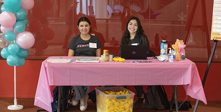 Two female students sit at a welcome table outside of the Women's Herstory Month event surrounded by balloons.