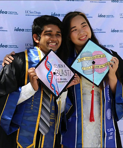 Two students in Commencement regalia hold up decorated caps