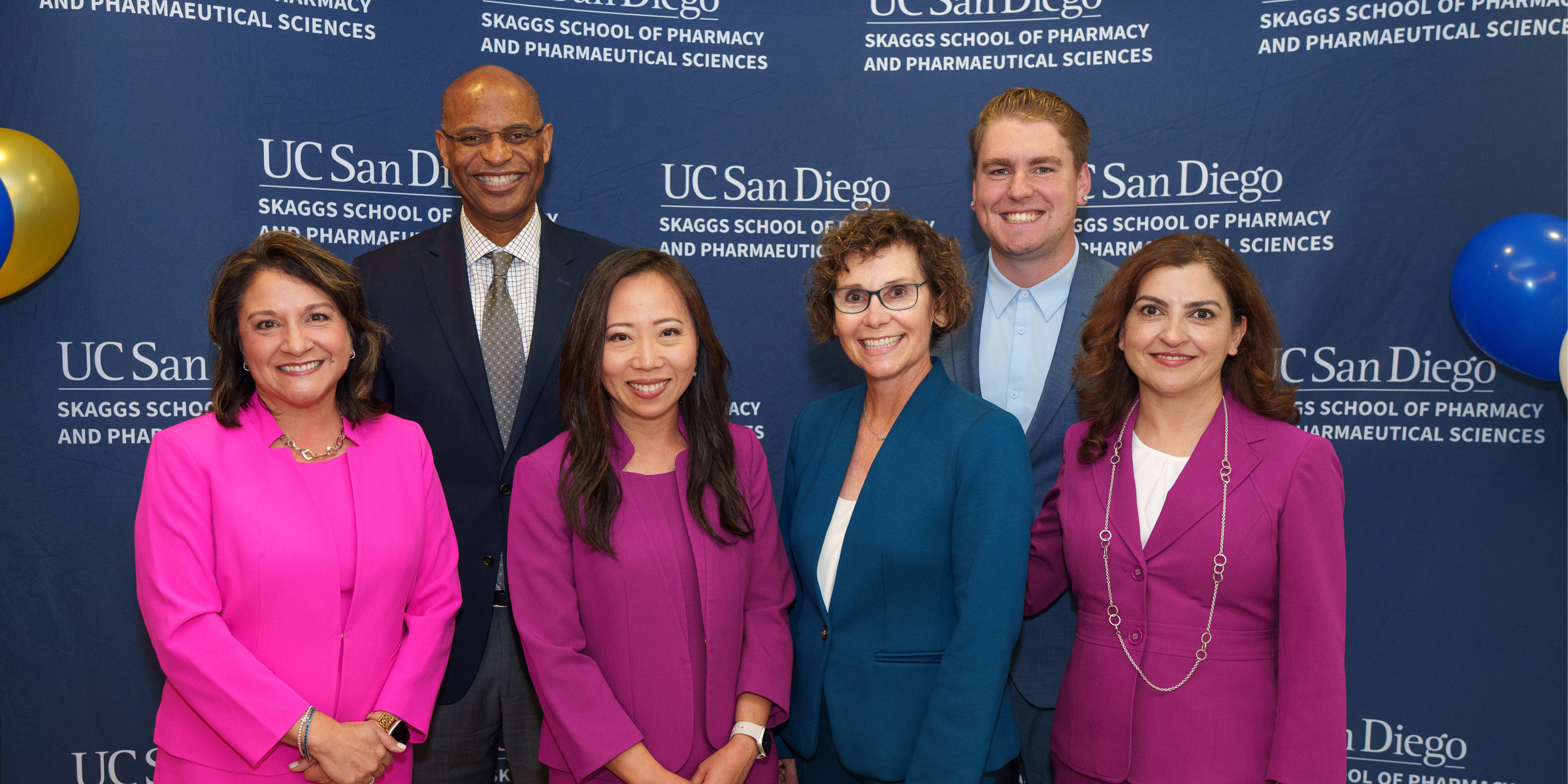 A group of six adults, two men (back) and three women, stand in front of a UC San Diego Skaggs School of Pharmacy and Pharmaceutical Sciences backdrop.