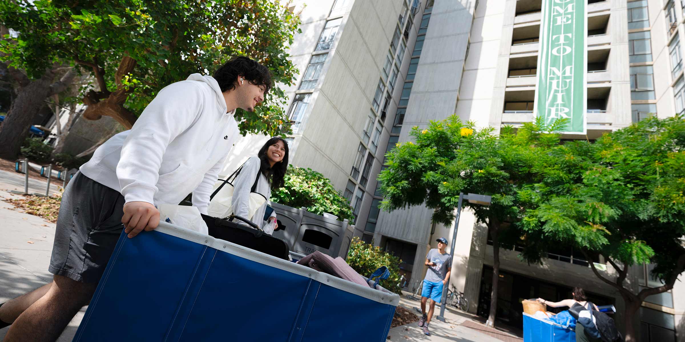 Two students wheel a cart full of luggage in front of residence halls