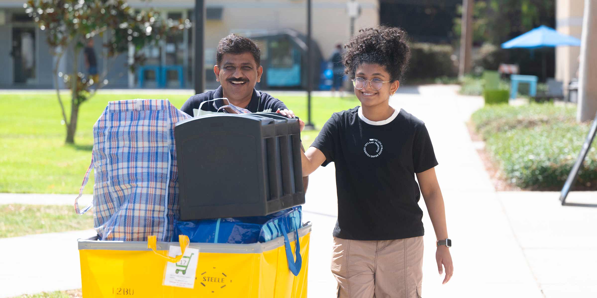 A student and an adult wheel a cart piled high with luggage.