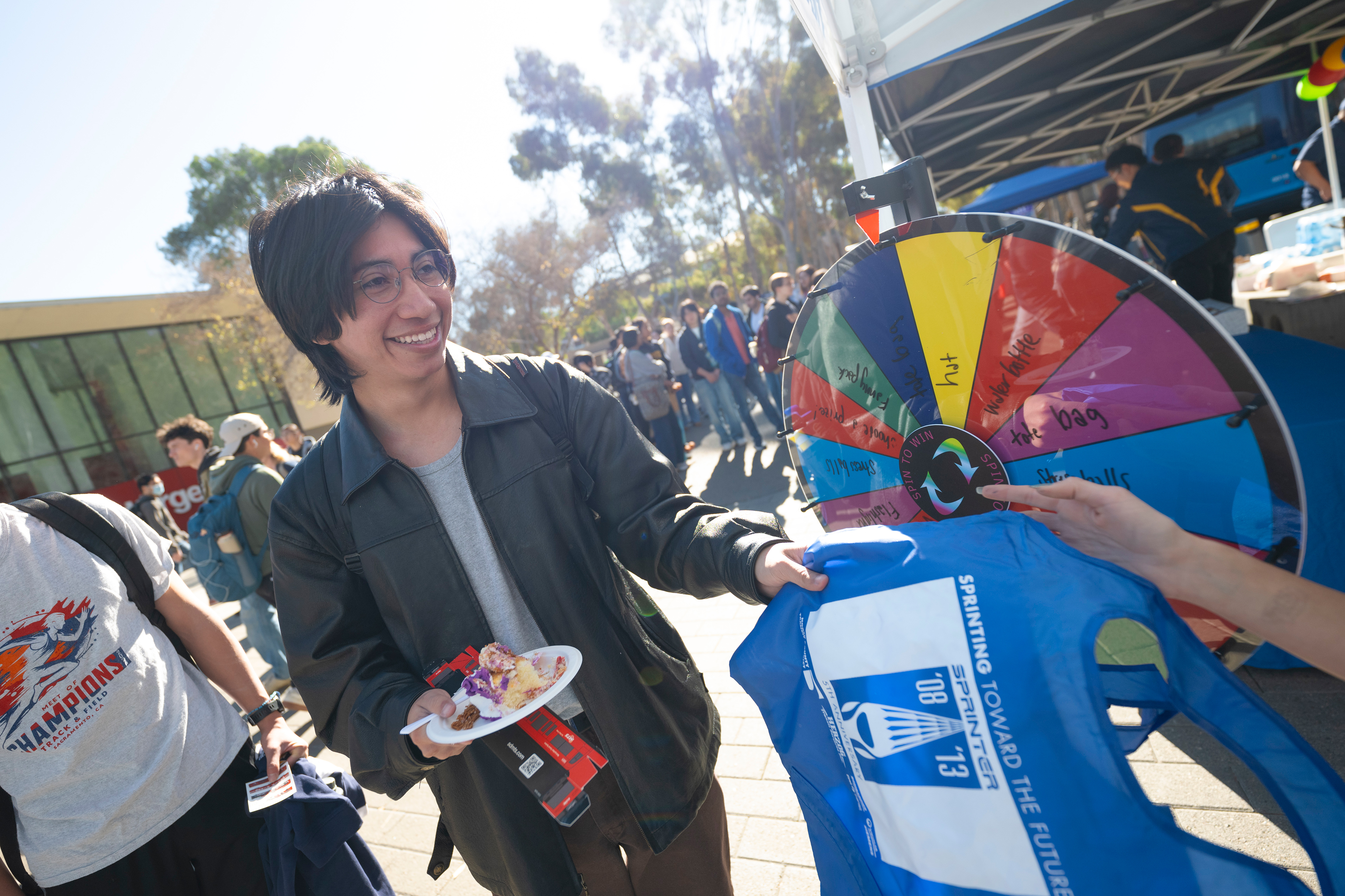 A person carrying a plate of a slice of cake grabs a blue branded bag from someone behind a booth