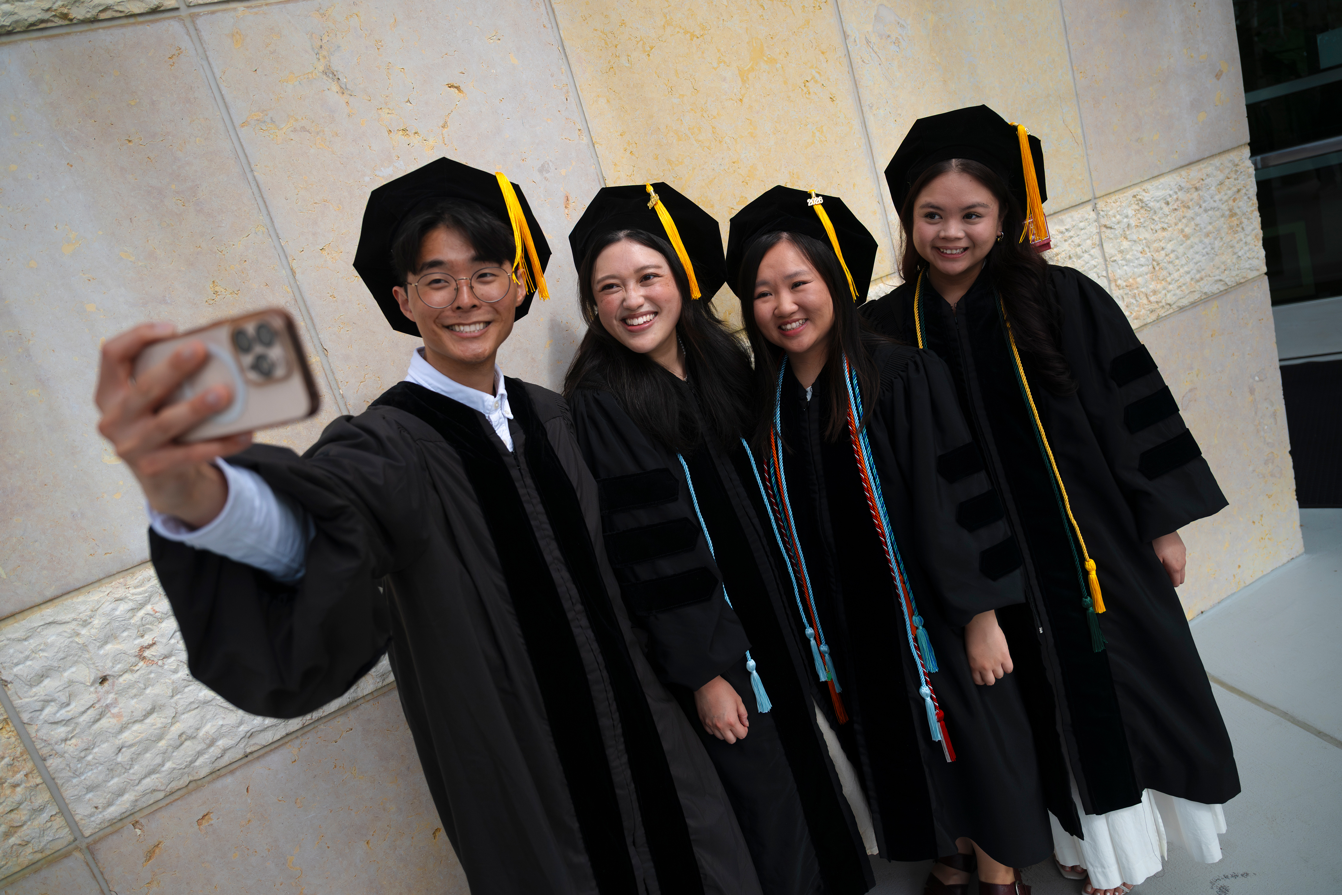 Male student holds phone while taking a selfie with colleagues in graduation gowns.