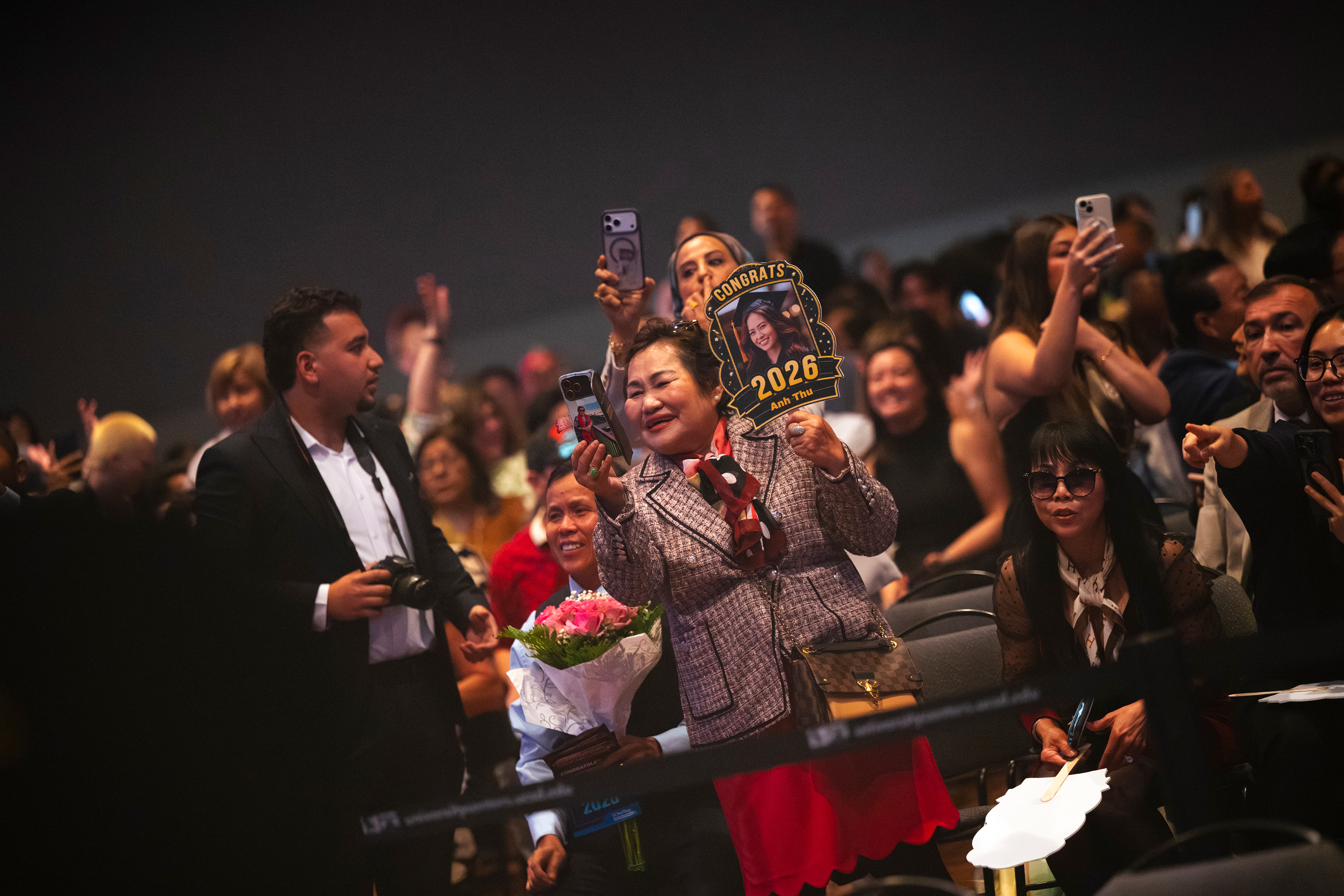 A mom holds a poster displaying a photo of her daughter, trying to take pictures during the graduation ceremony.