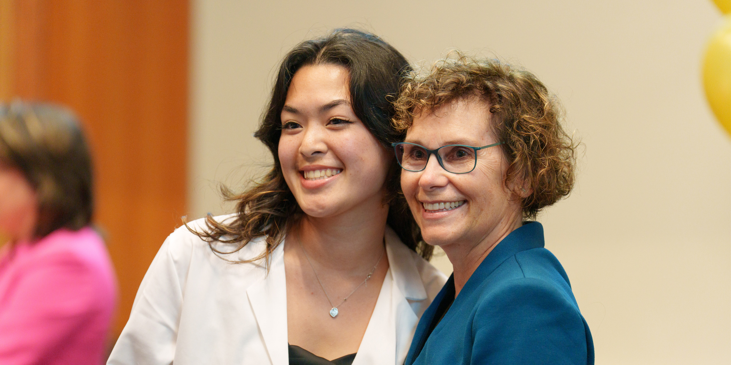 Two people pose together at an event, female on the left is wearing white coat and woman on the right is wearing a blue blazer jacket.
