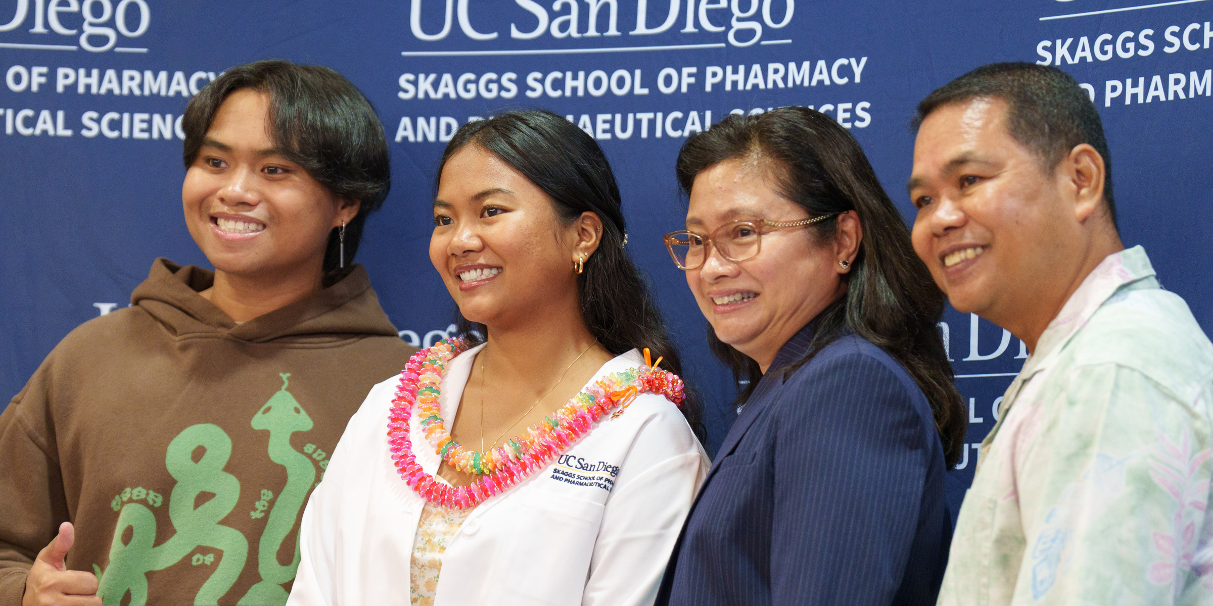 Four people posing for the camera with the UC San Diego Skaggs banner. Main female is in center with white coat jacket and leis.