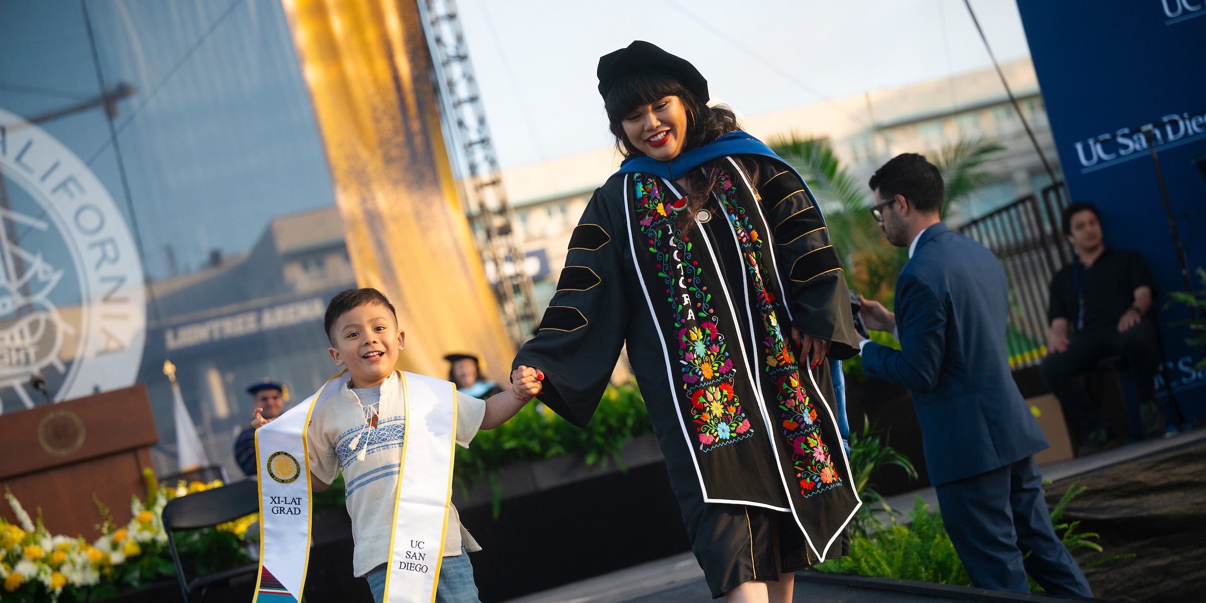 A graduate walking across the stage holding hands with a young child
