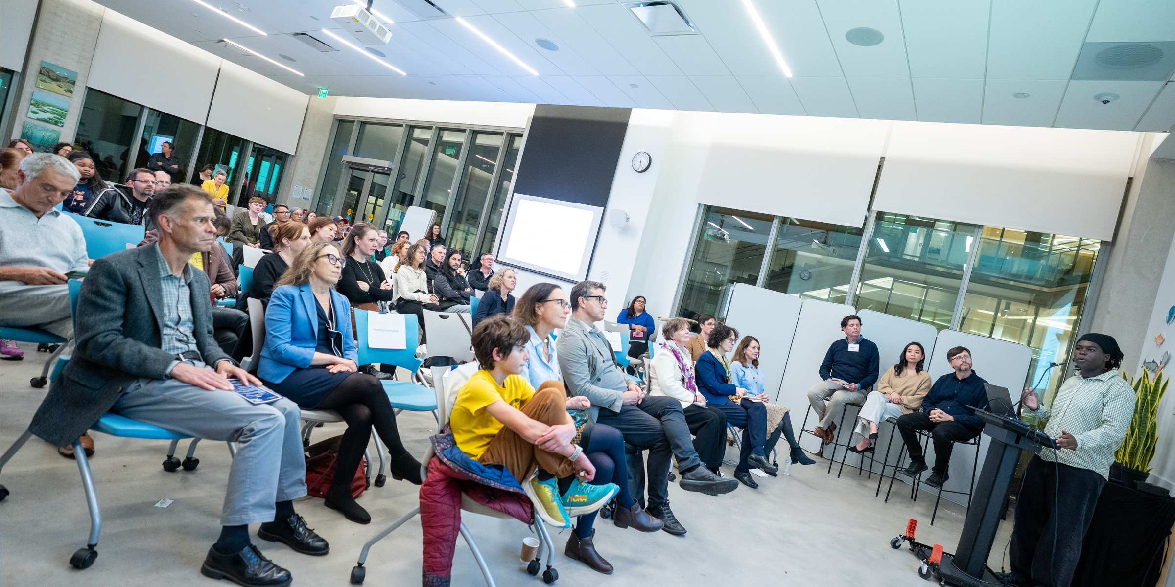 A group of people listening to a formal talk by a student