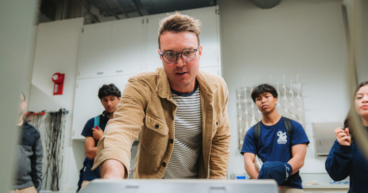 Slideshow: scientist in brown jacket talks to students in lab