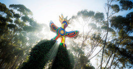 The sun shines behind a colorful bird statue surrounded by trees. 