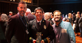 Photo of three smiling men in a crowded room, with the middle man in a uniform and holding a glass award.