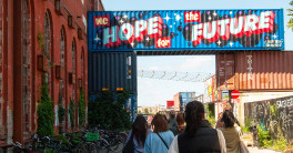 A group of people walk into an outdoor market built of shipping containers. One of the containers is painted with a mural that says 