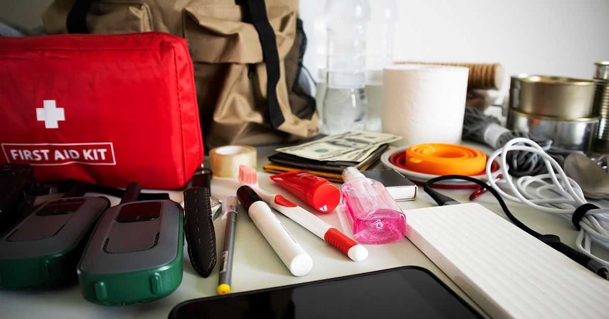 A backpack with emergency supplies such as phone charger, first aid kit, phone cord, canned food and water bottles sits on a table. 