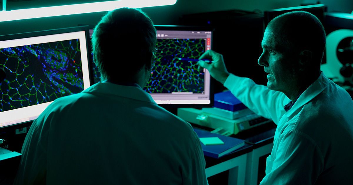In a room lit with a blue hue, two researchers look at imaging on computer monitors.