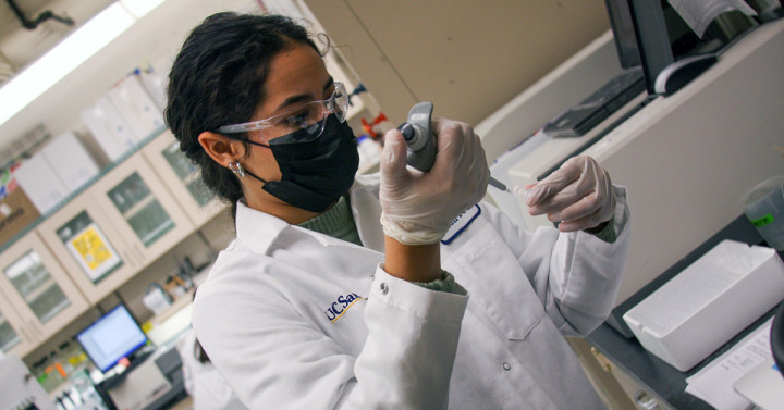 A student wearing a white lab coat uses a pipette to put samples in a test tube.