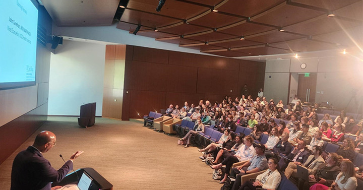 Man standing at the podium in a large auditorium speaking to a group of people. 
