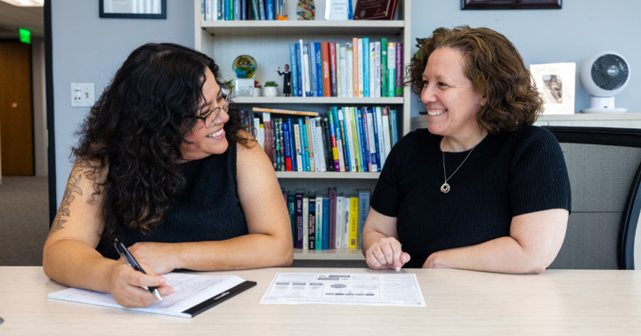 Two women sitting at a table with a notebook and other papers. They are looking at each other and smiling.