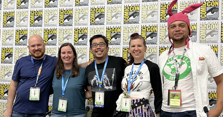 A group of five people three men and two women with their arms around one another posing and smiling in front of a Comic-Con banner. 