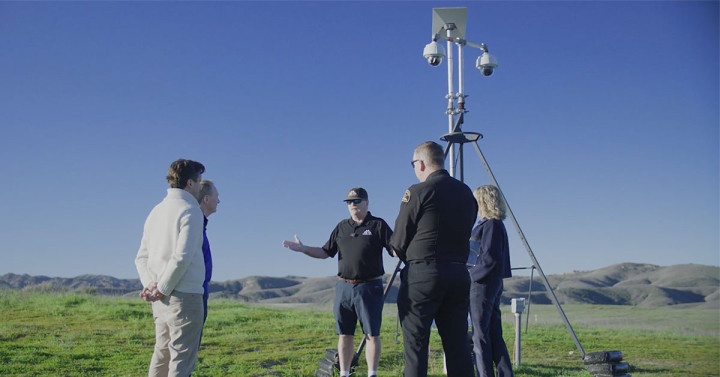 Five people stand on a grassy hill on a sunny day in front of an ALERTCalifornia tripod camera unit. The tripod has two bowling ball sized cameras attached. 