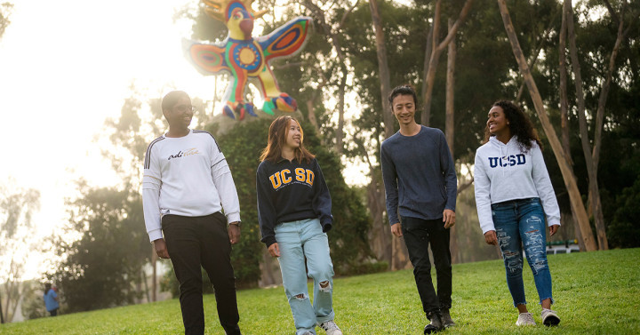 Four smiling students walk across a lawn with a large, colorful bird sculpture behind them. Two students are wearing jackets that say 