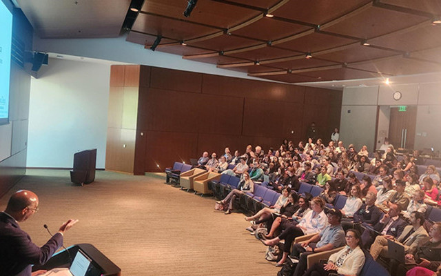 Man standing at the podium in a large auditorium speaking to a group of people. 