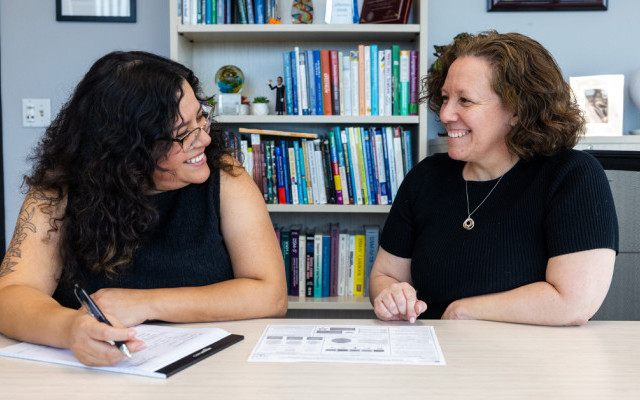 Two women sitting at a table with a notebook and other papers. They are looking at each other and smiling.
